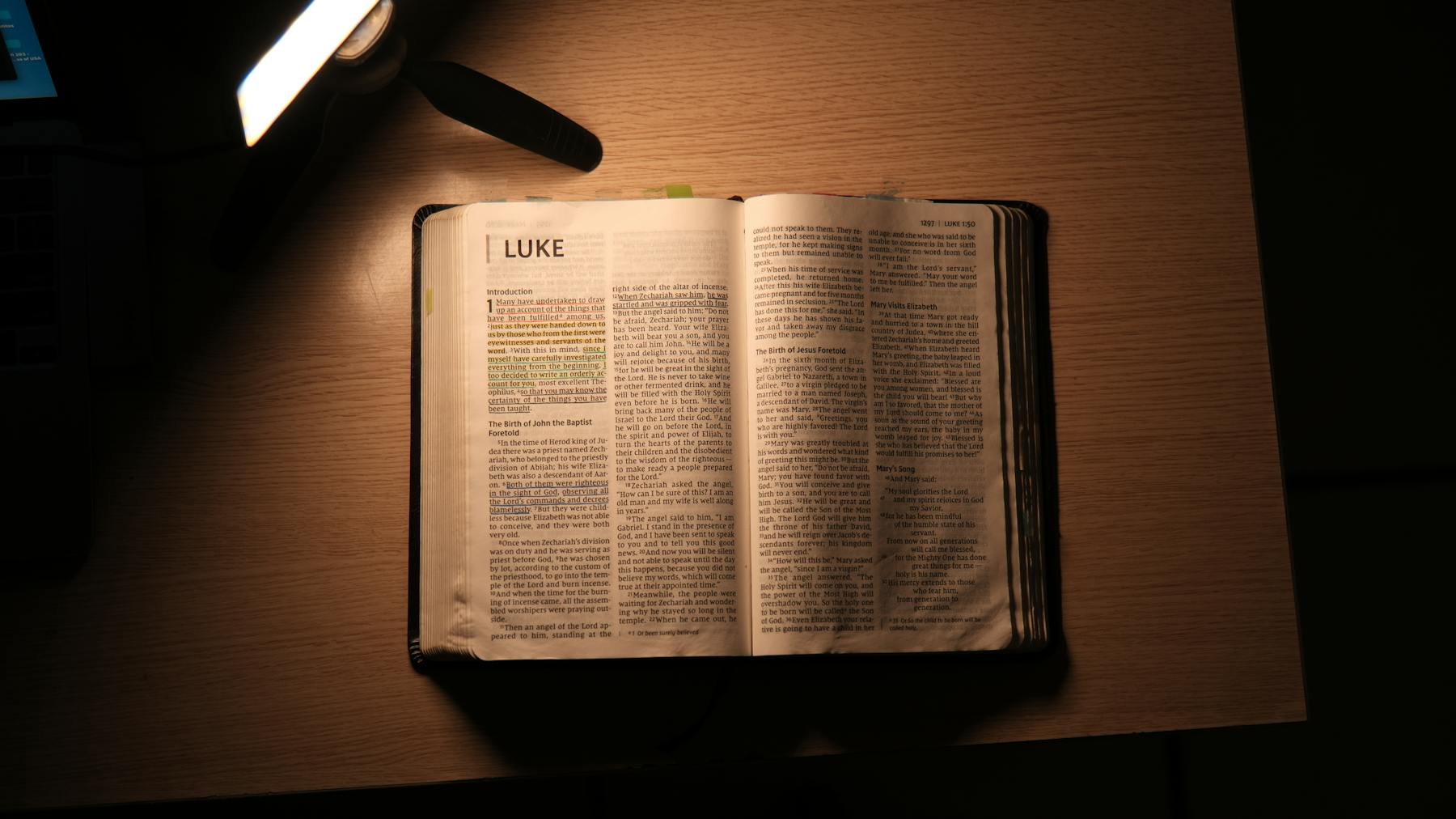An open Bible on a wooden desk under warm lamp light.